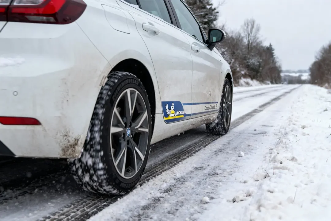 Voiture blanche roule sur route enneigée, équipée pneus Michelin CrossClimate 2, paysage hivernal en arrière-plan.
