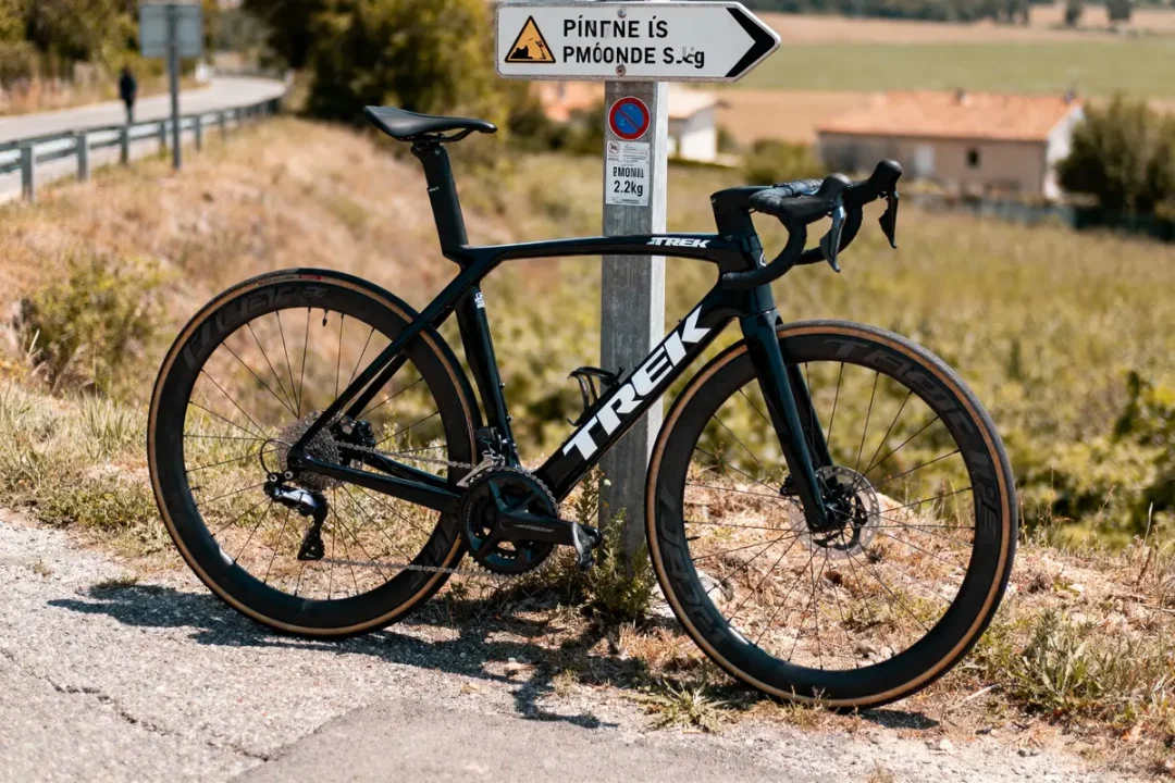 Vélo Trek noir stationné près d'un panneau directionnel sur route ensoleillée. Paysage champêtre en arrière-plan.