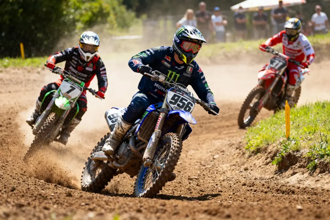 Trois motocyclistes en course sur terrain poussiéreux, en pleine accélération, portant des tenues de compétition.
