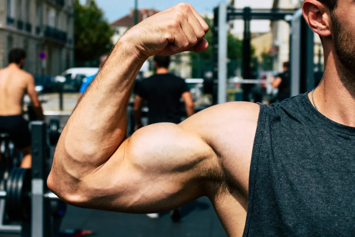 Bras musclé fléchissant à l'extérieur, focus sur biceps. En arrière-plan, personnes pratiquant la musculation.