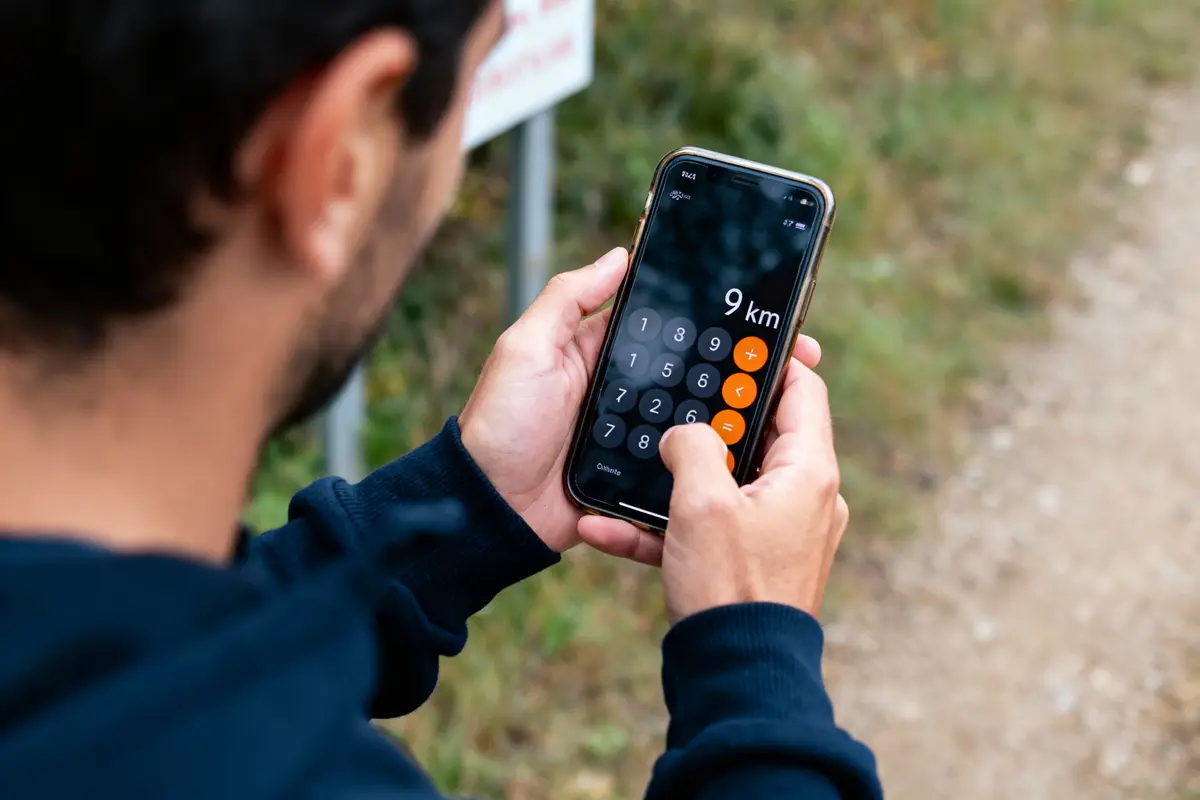Personne utilisant une calculatrice sur un smartphone affichant "9 km", en extérieur, portant une veste bleue foncée.
