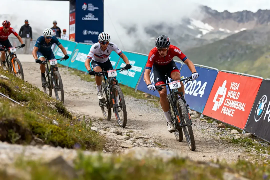 Quatre cyclistes en compétition sur un sentier montagneux lors d'une course mondiale en France. Panneaux publicitaires présents.