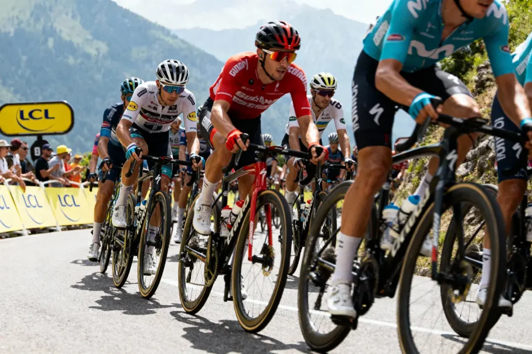 Cyclistes en peloton lors d'une course de montagne, spectateurs en arrière-plan, ciel partiellement nuageux.