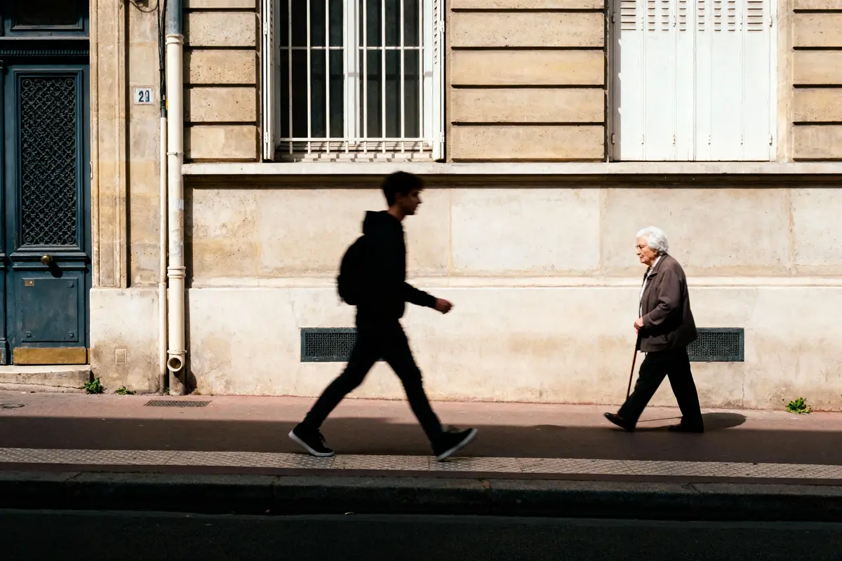 Deux personnes marchent sur le trottoir, devant un mur beige, éclairées par le soleil.