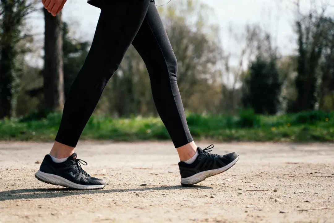 Personne marchant sur chemin de terre, portant leggings noirs et baskets. Arrière-plan flou avec arbres et verdure.