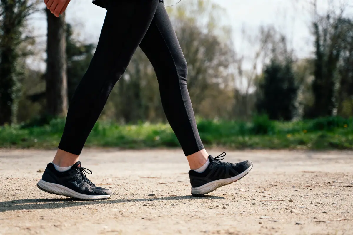 Personne marchant sur chemin de terre, portant leggings noirs et baskets. Arrière-plan flou avec arbres et verdure.