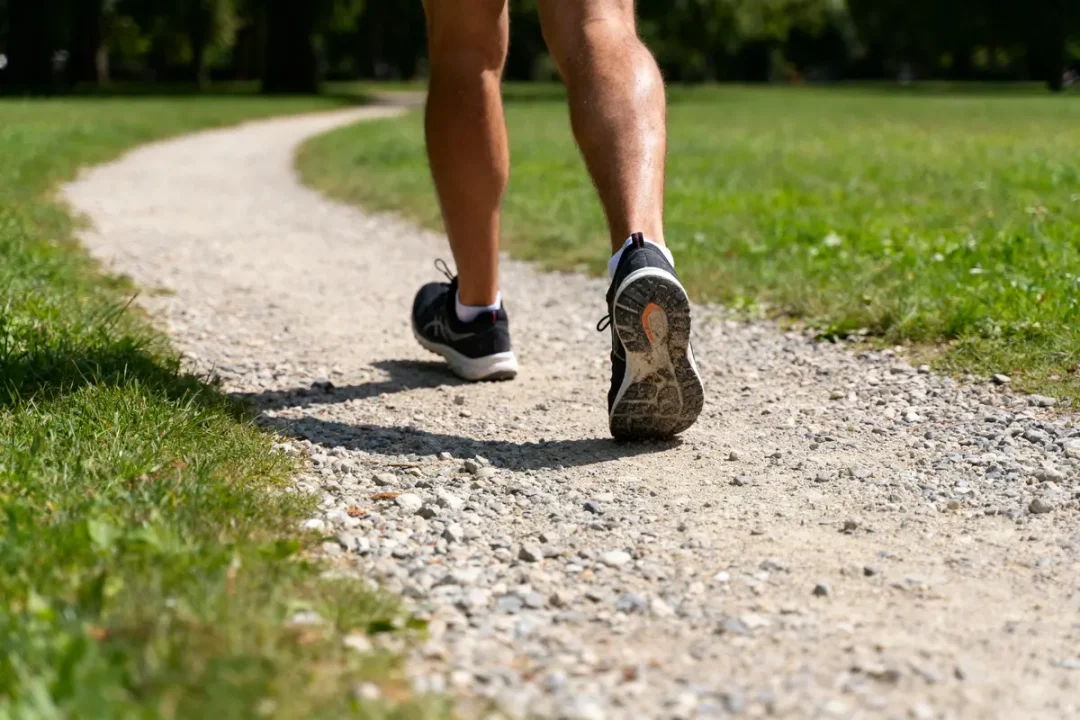 Jambes d'une personne marchant sur chemin gravillonné dans un parc, portant des baskets noires.