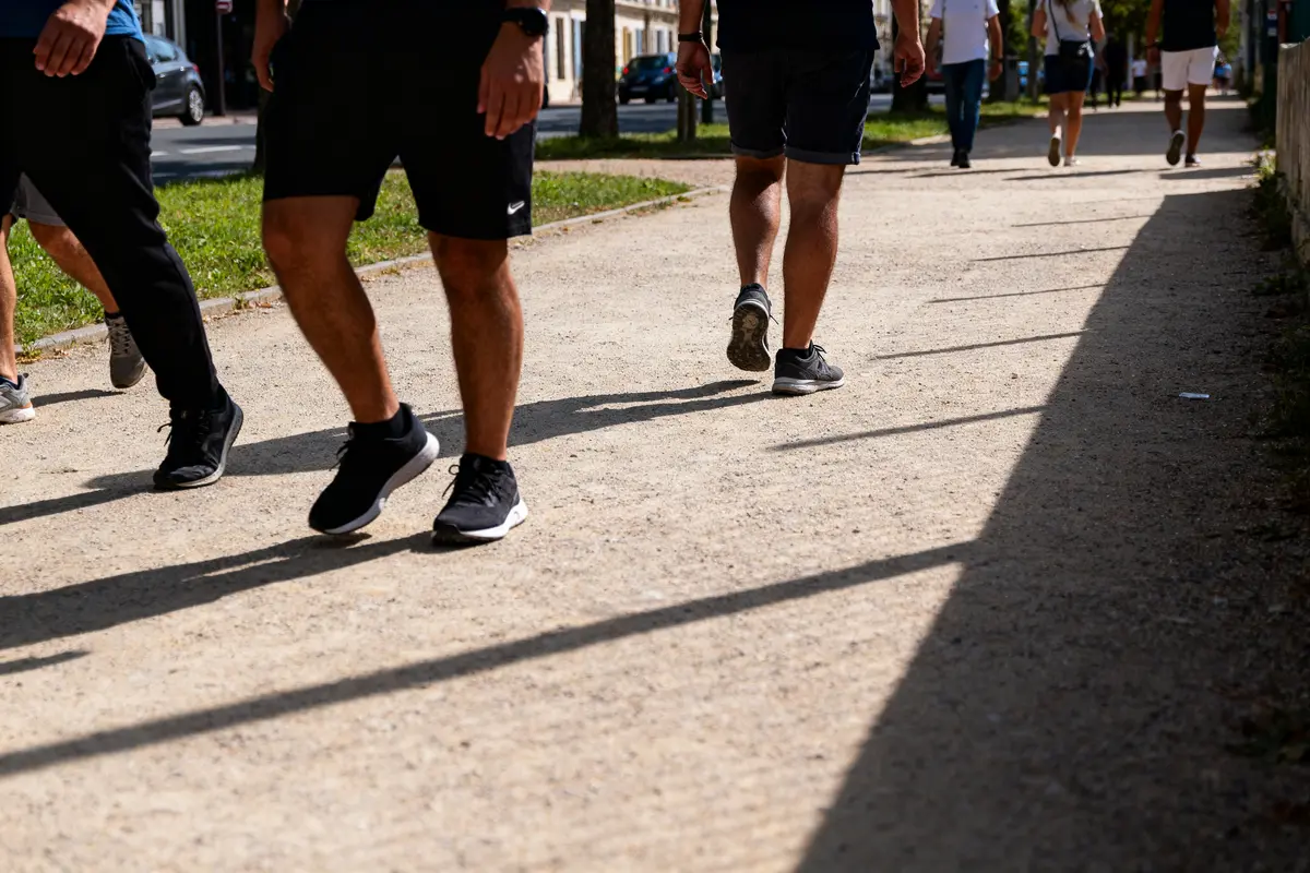 Personnes marchant sur un chemin ensoleillé, portant des vêtements sportifs et baskets noires. Ombres projetées au sol.