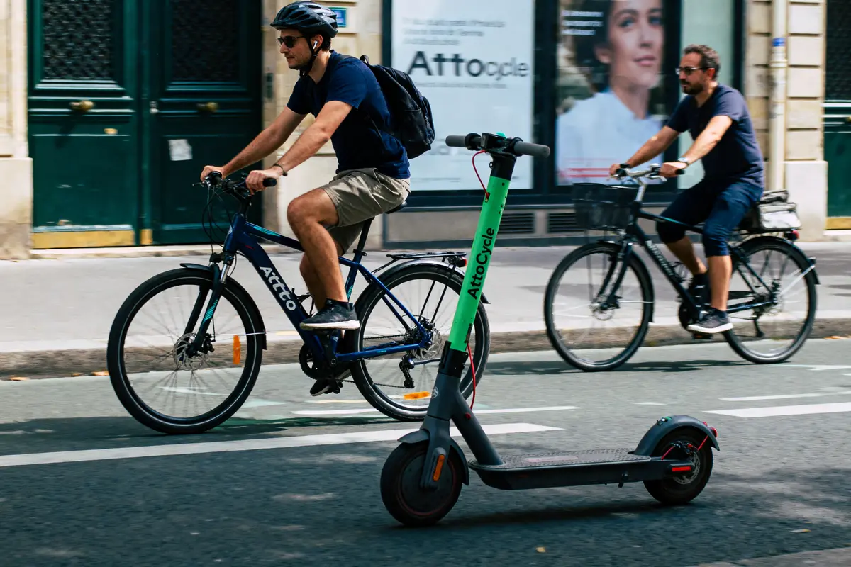 Deux cyclistes roulent en ville, passant à côté d'une trottinette verte AttoCycle stationnée.
