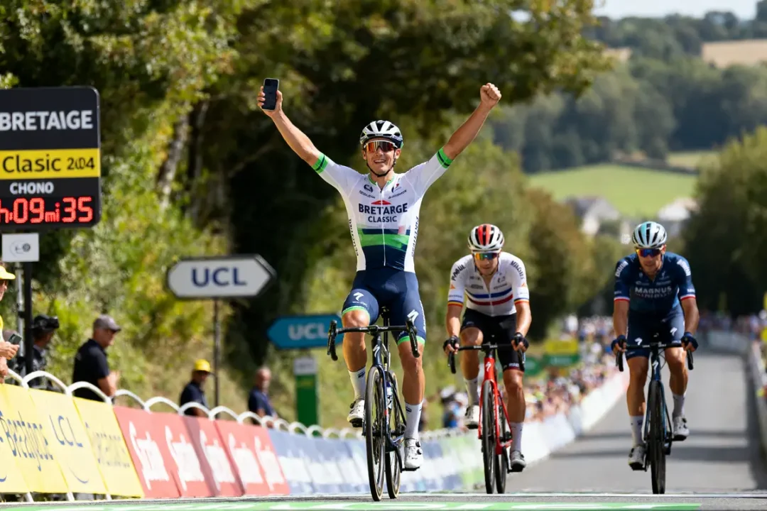 Cycliste levant les bras en signe de victoire sur la ligne d'arrivée d'une course, deux concurrents derrière.