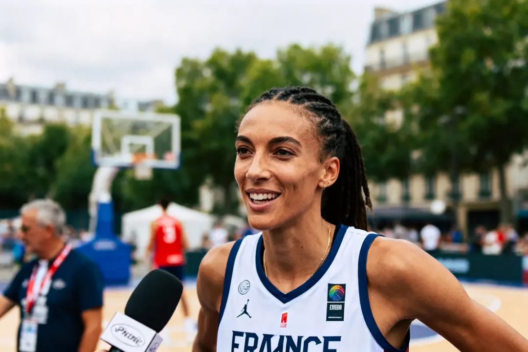 Joueuse de basket interviewée, portant maillot France, extérieur, terrain avec panier et spectateurs en arrière-plan.