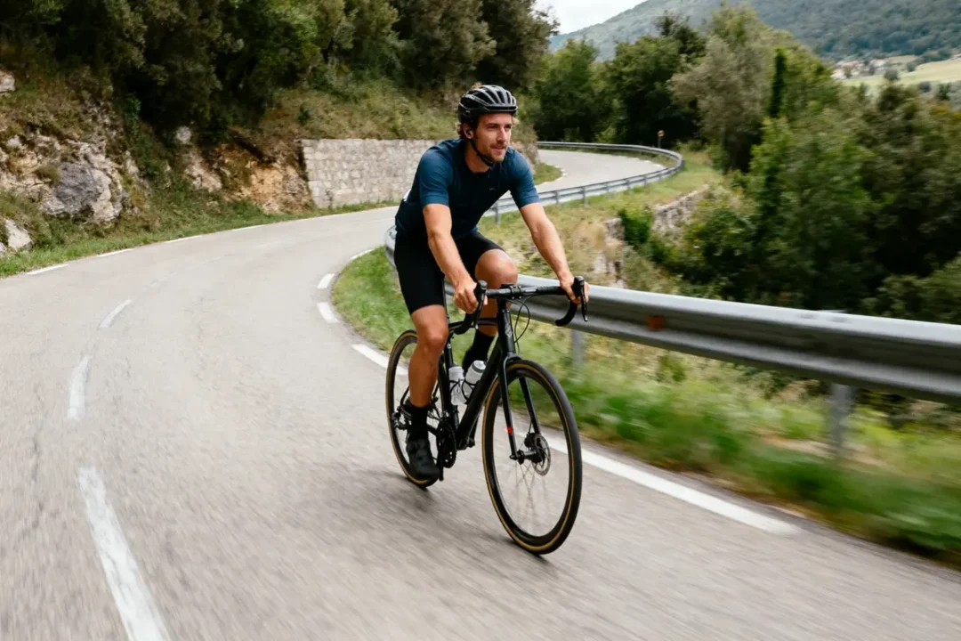 Cycliste en montagne sur route sinueuse, vêtu de noir, portant casque, entouré de forêts verdoyantes.