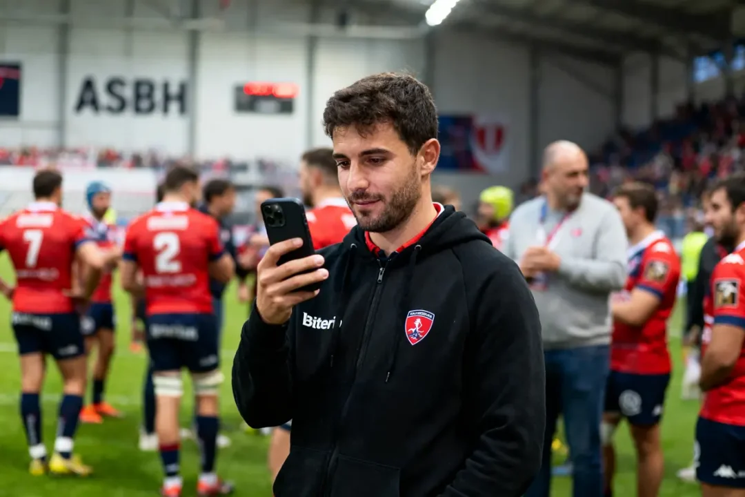 Homme lisant sur smartphone, en sweat noir, devant joueurs de rugby en tenues rouges, intérieur stade.