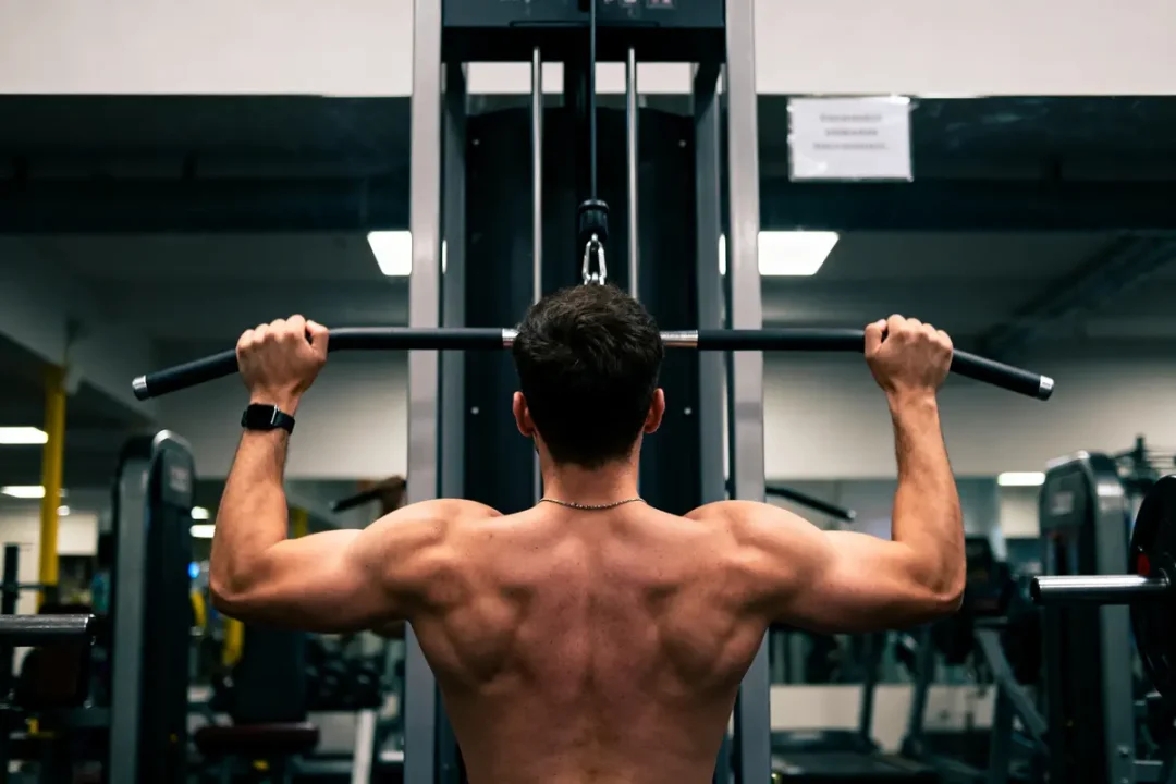 Homme musclé de dos, fait des tractions à la barre dans une salle de sport moderne.