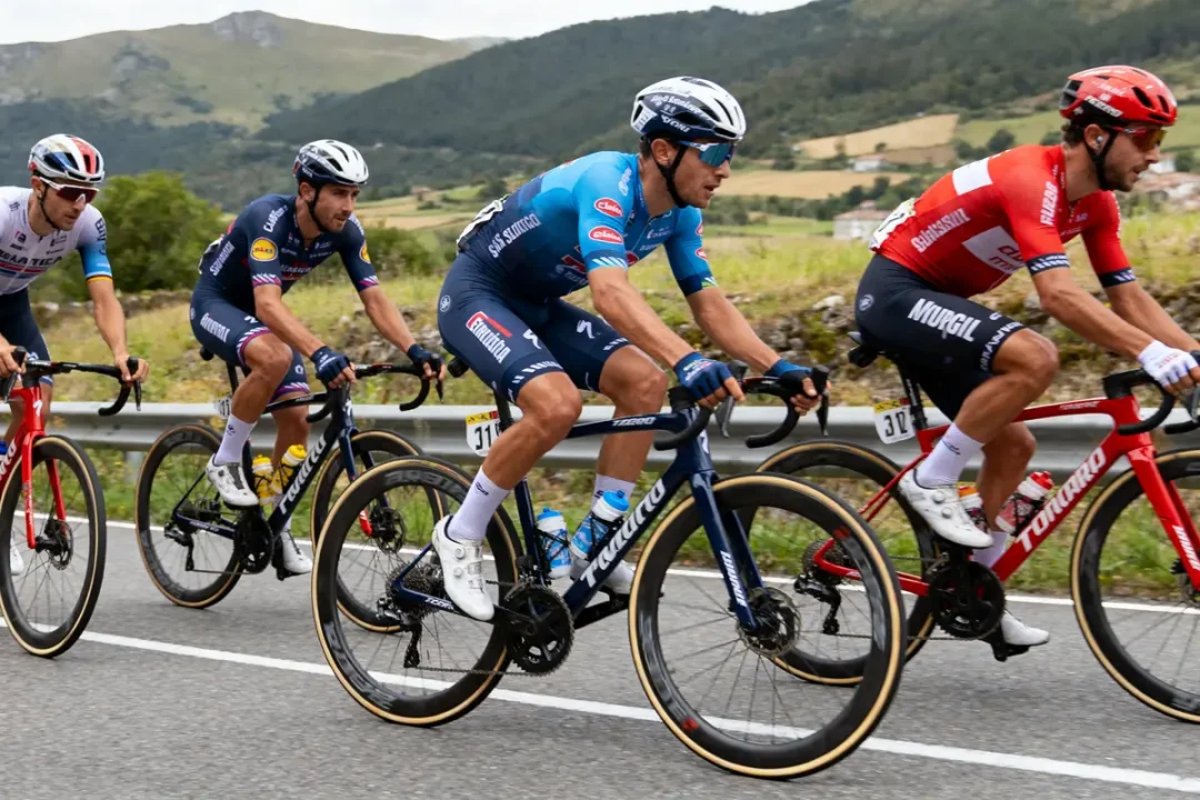 Quatre cyclistes professionnels en course, pédalent sur une route de campagne, montagnes en arrière-plan.