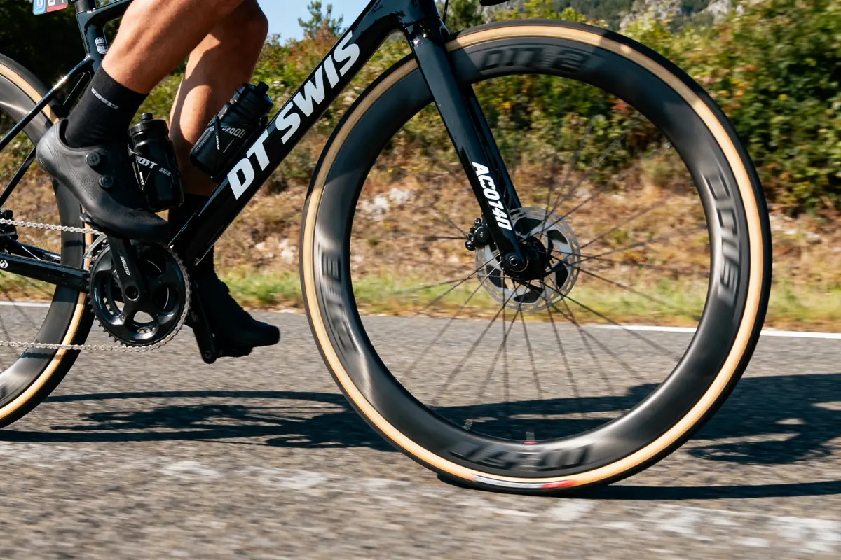 Cycliste sur route, pédalant, vélo DT Swiss noir, gros plan sur la roue et les jambes, environnement naturel.