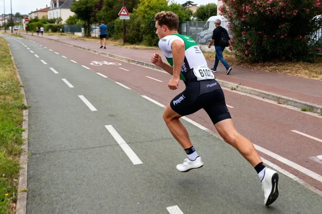 Homme courant sur une piste cyclable, vêtu de vêtements de sport, avec dossard, arbres et personnes en arrière-plan.