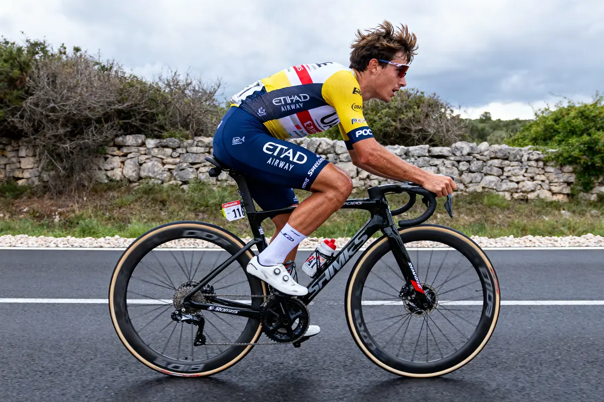 Coureur cycliste en action sur route, portant un uniforme jaune et bleu, vélo noir, nature en arrière-plan.