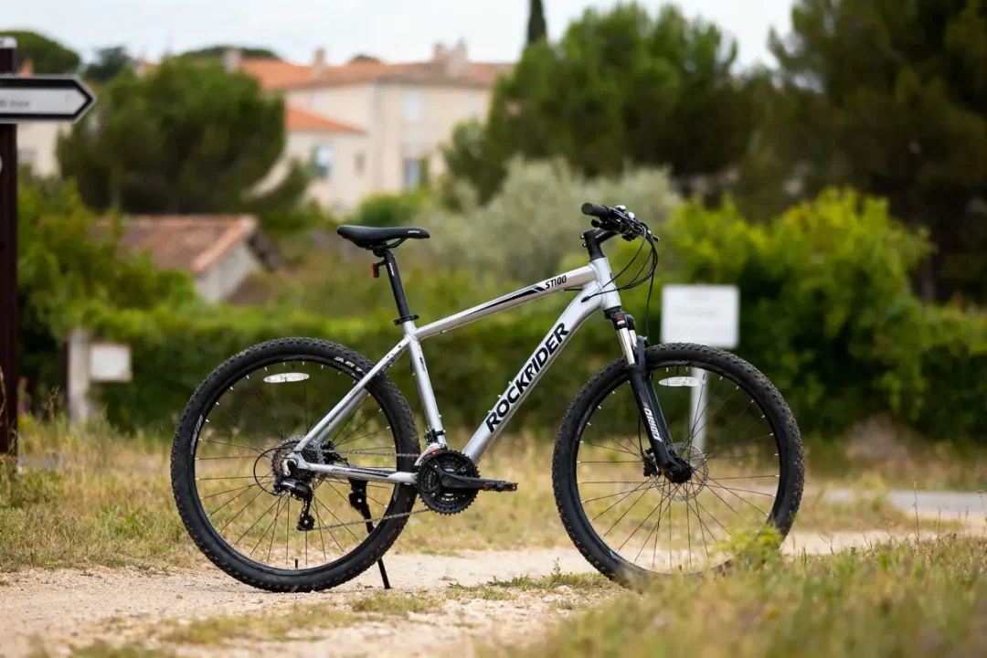 Vélo de montagne argenté Rockrider ST100 sur chemin, végétation en arrière-plan.