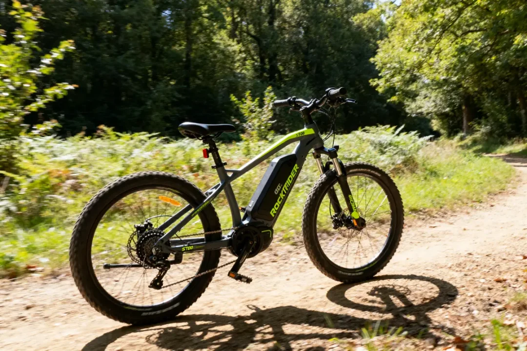 Vélo tout-terrain électrique Rockrider ST100 sur sentier de forêt ensoleillé, à côté de la végétation verte.