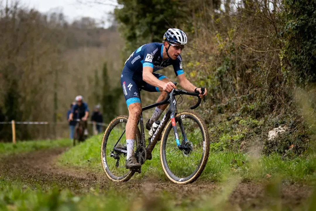 Cycliste en course sur un chemin boueux, portant un uniforme bleu et casque, entouré d'arbres.