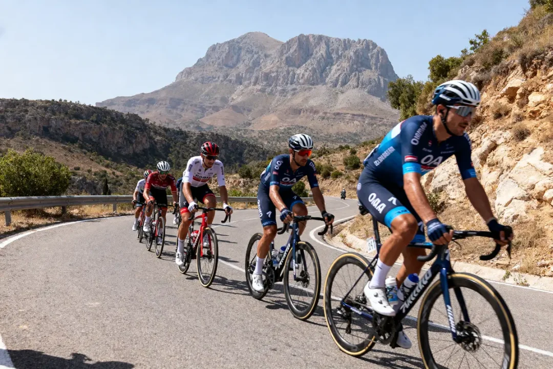 Cyclistes en course sur une route montagneuse, entourés de paysages rocheux et verdoyants, sous un ciel dégagé.