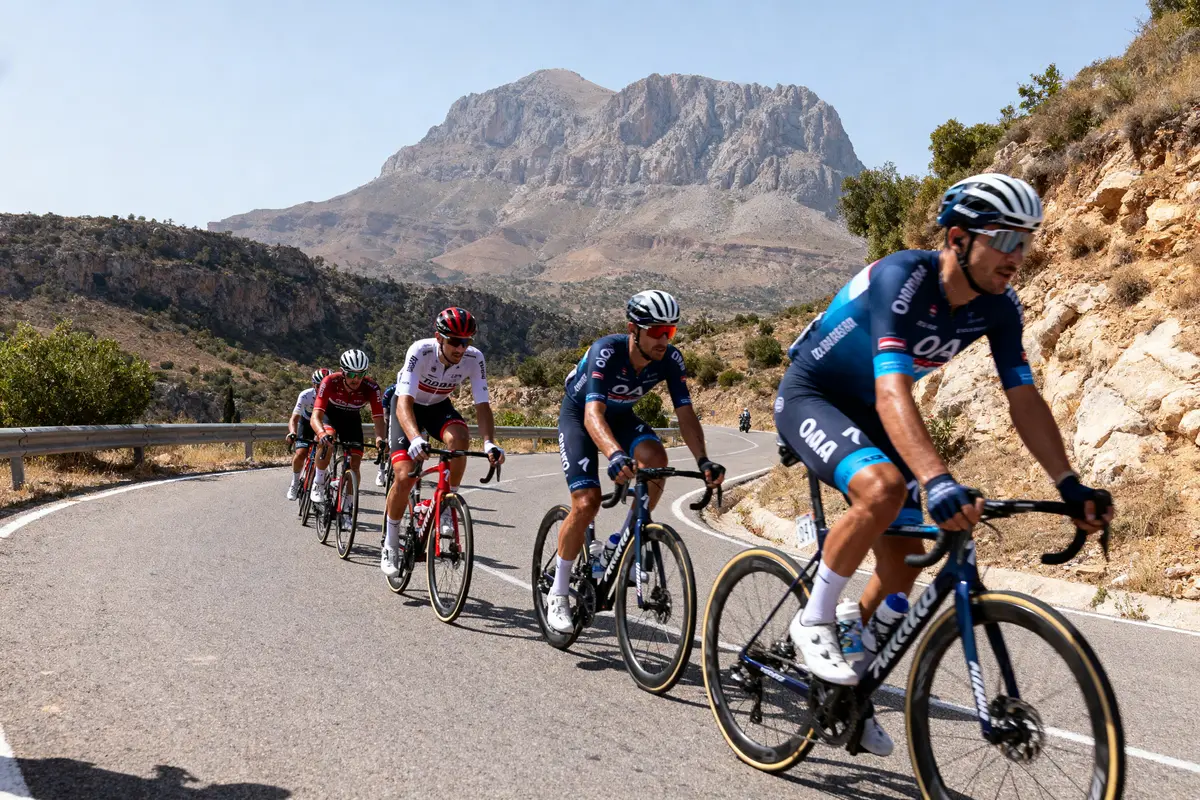 Cyclistes en course sur une route montagneuse, entourés de paysages rocheux et verdoyants, sous un ciel dégagé.