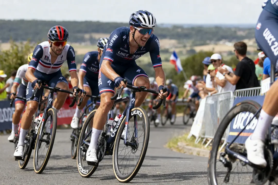 Des cyclistes en uniforme bleu en course, avec spectateurs et drapeau français en arrière-plan.