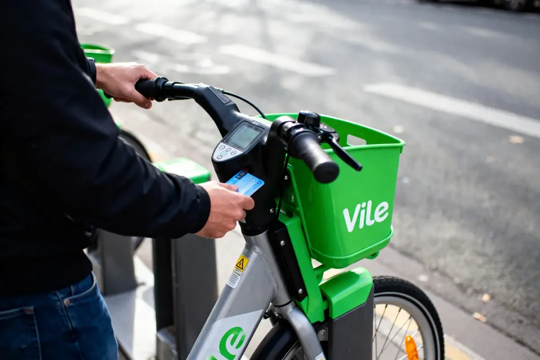 Personne activant un vélo en libre-service vert avec carte, station de location en arrière-plan.