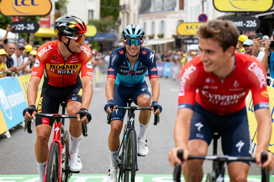 Trois cyclistes en compétition sur route, souriant, entourés de spectateurs, avec arrière-plan urbain.
