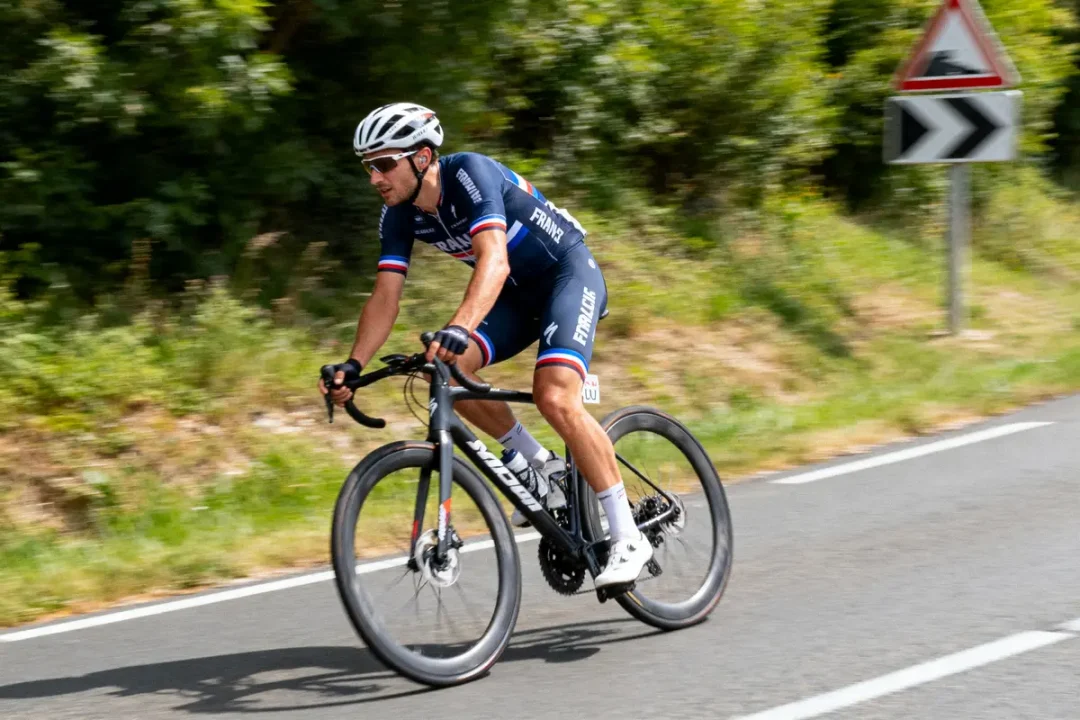Cycliste en tenue de France sur route, forêt environnante, panneau de virage à droite visible en arrière-plan.