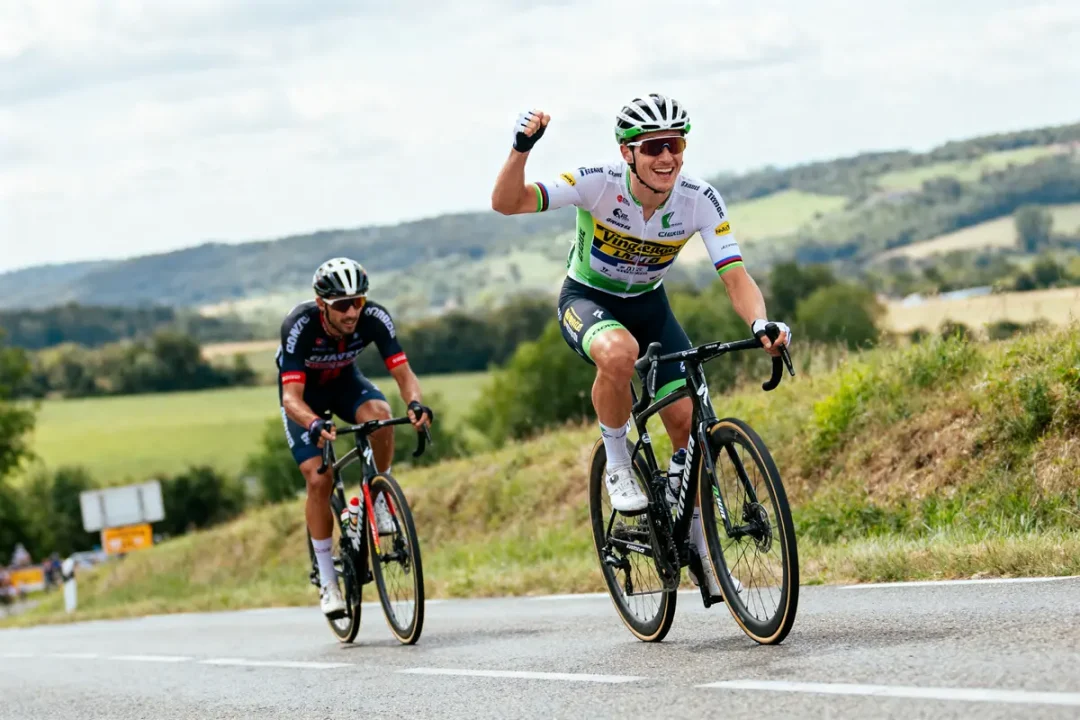Deux cyclistes en compétition, l'un levant le poing, sur une route de campagne vallonnée.