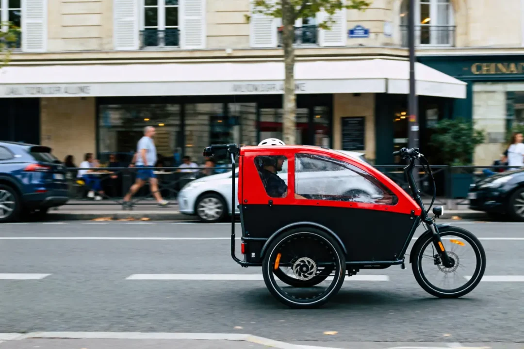 Triporteur électrique rouge et noir circulant en ville, avec passager portant un casque, devant des bâtiments.