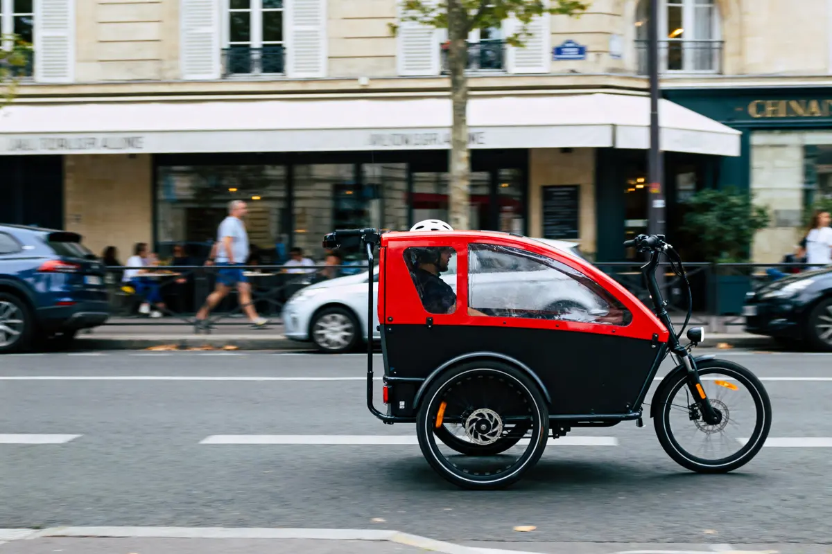 Triporteur électrique rouge et noir circulant en ville, avec passager portant un casque, devant des bâtiments.