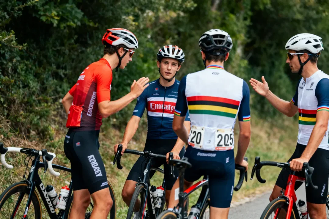 Quatre cyclistes discutent ensemble en tenue de course, sur une route bordée de verdure.