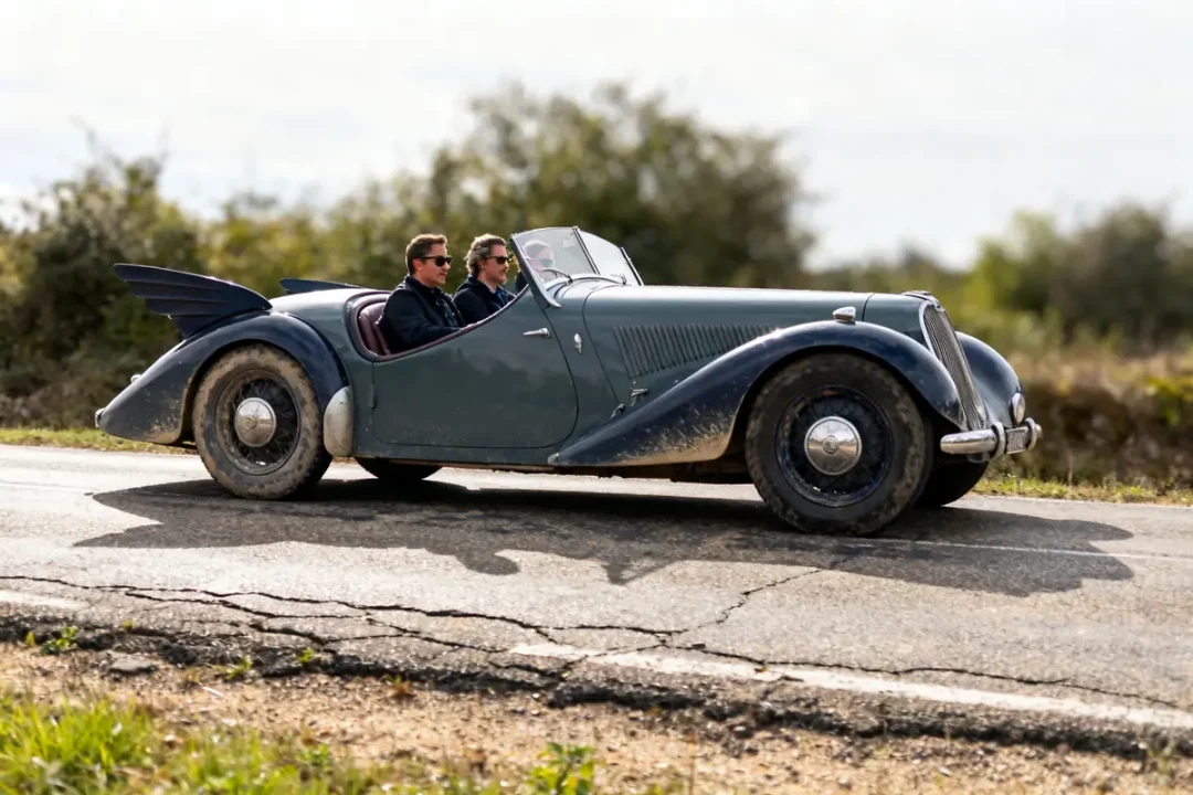 Deux personnes conduisent une Renault Vivastella Grand Sport historique sur une route campagne.