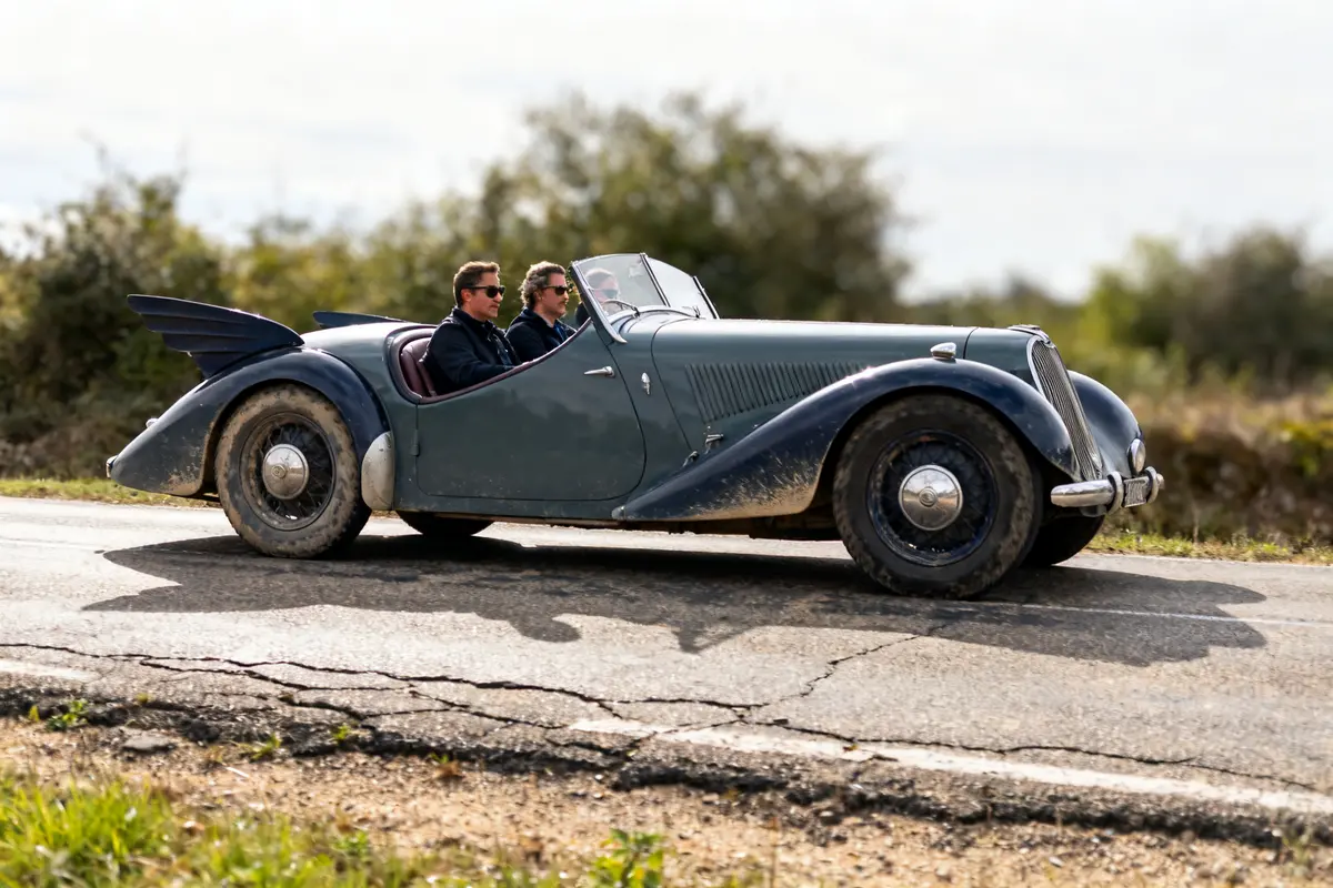 Deux personnes conduisent une Renault Vivastella Grand Sport historique sur une route campagne.