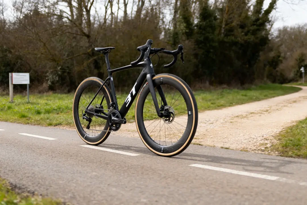 Vélo de route noir 3T Exploro Racemax sur piste cyclable bordée d'arbres.